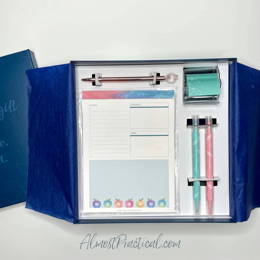 box with teacher planning pad, rose gold ballpoint pen with apple design on end, 2 highlighters, roll of sticky note tape.