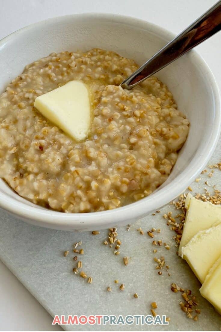 steel cut oatmeal in a white bowl with a pat of butter in center with raw oats sprinkled around the bowl and some extra pats of butter