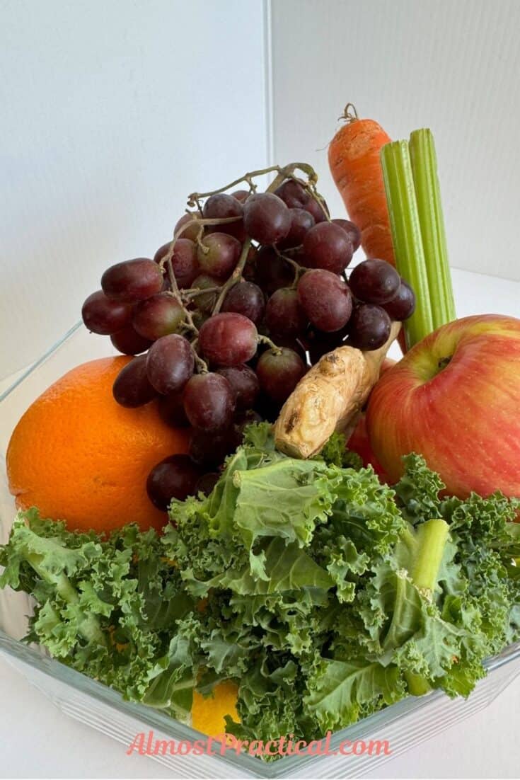 a collection of fruits and vegetables in a glass bowl including red grapes, orange, ginger, kale, apple, celery, and carrot